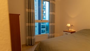 Un dormitorio con cama, mesita de noche con lámpara, ventana con cortinas y un armario de madera.