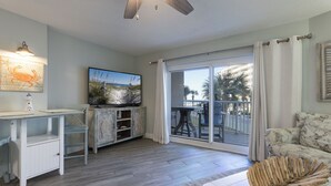 A spacious living room with a large TV, a wooden coffee table, and a view of the outdoors through a sliding glass door. - Tradewinds 108- 1 Bedroom with Hall Bunks! 2 Full Bathrooms! Updated! (Orange Beach)