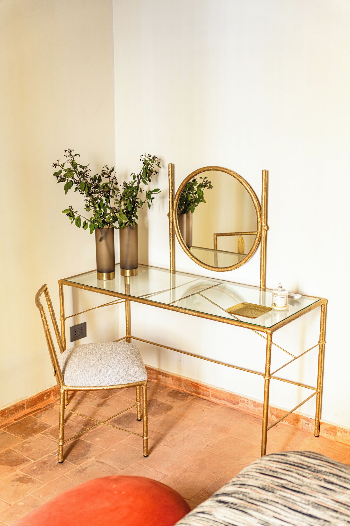 Une coiffeuse en verre avec un cadre doré, un miroir rond et deux vases avec des plantes.