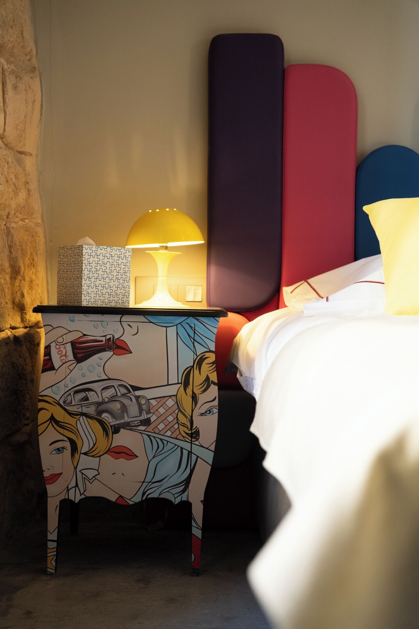 Un lit recouvert d’une couette blanche, une lampe de chevet et une table de chevet colorée au motif pop art.