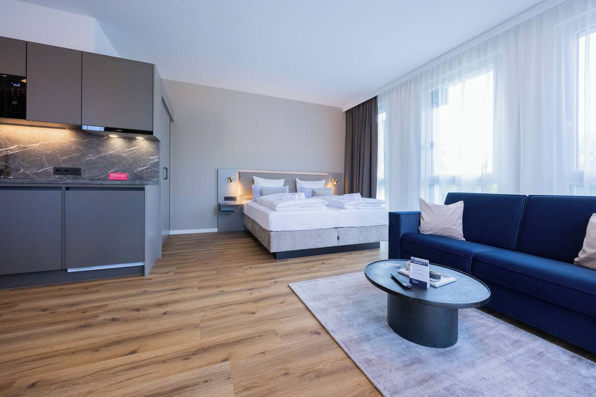 Een moderne hotelkamer met een kitchenette, een bed met witte lakens, een blauwe bank en een ronde salontafel.