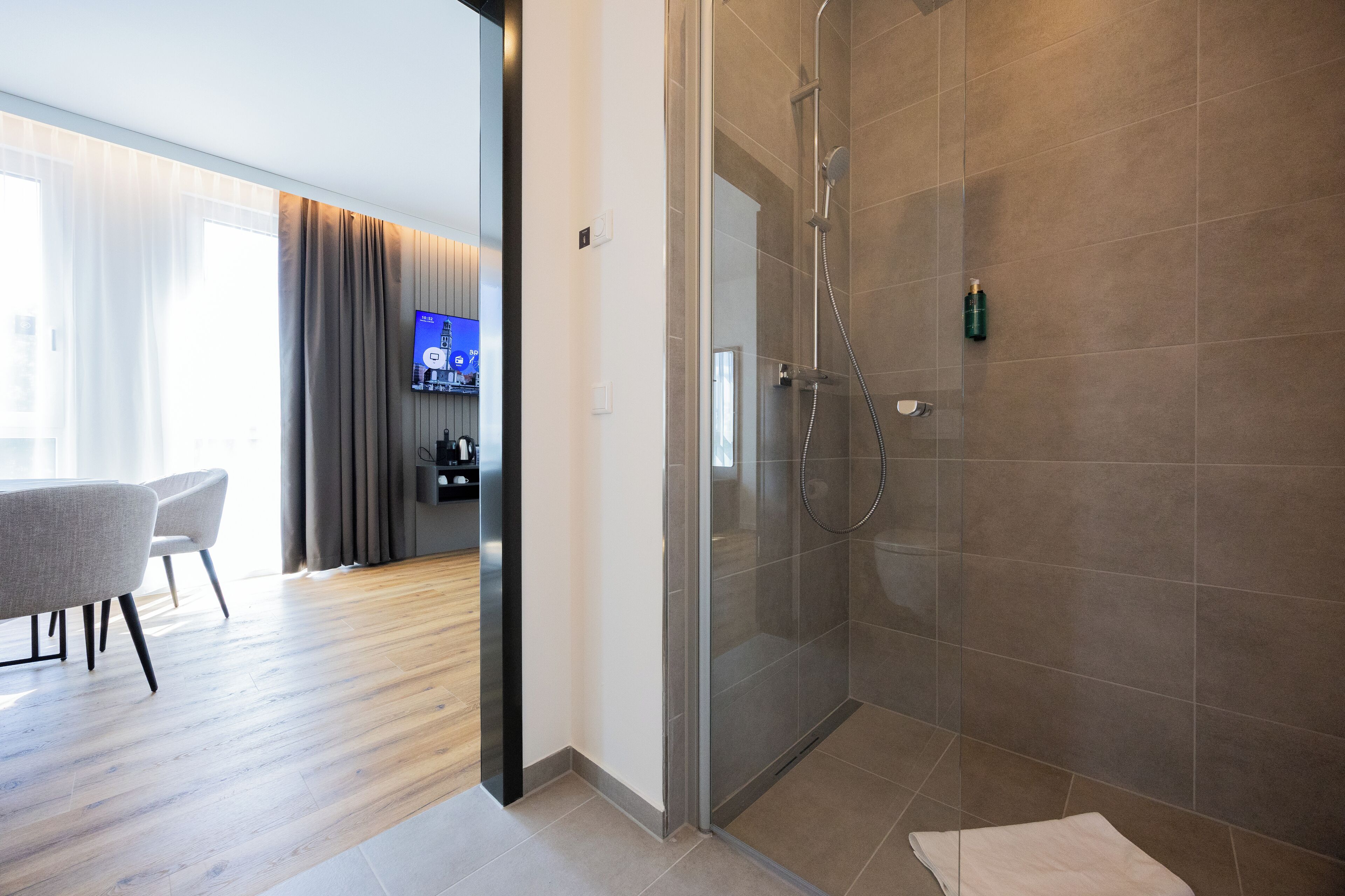 Una habitación de hotel moderna con ducha de vidrio, suelo de madera, zona de comedor con sillas y televisor de pantalla plana.