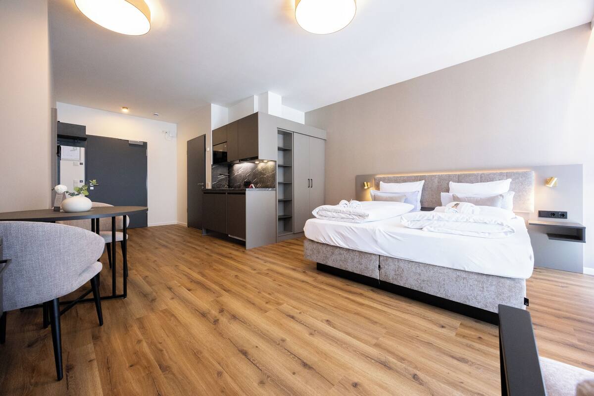 Een moderne hotelkamer met een houten vloer, een bed met witte lakens, een bureau met stoel en een geïntegreerde keukenhoek.