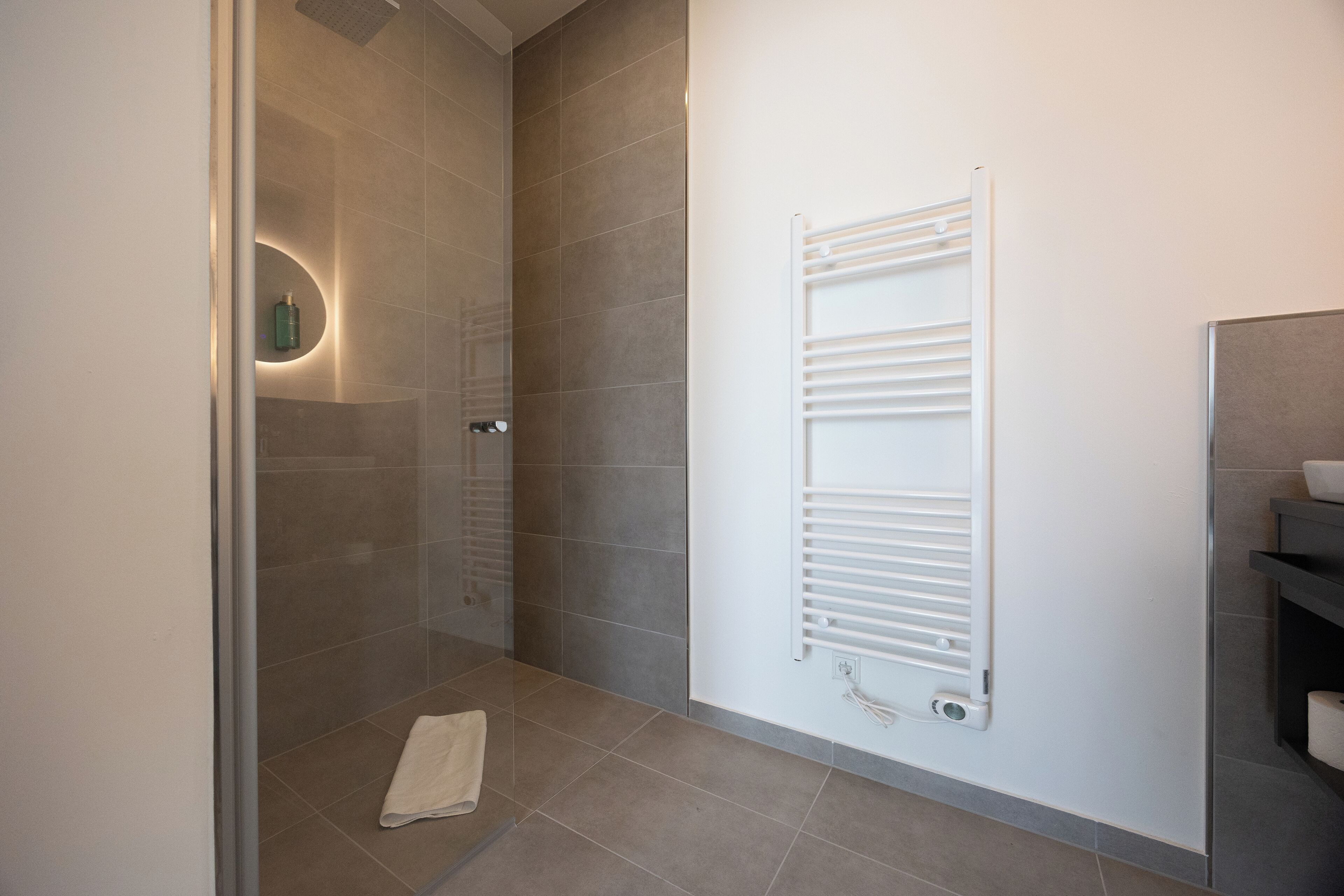 A modern bathroom with a glass shower enclosure, a white towel on the floor, and a wall-mounted towel rack.
