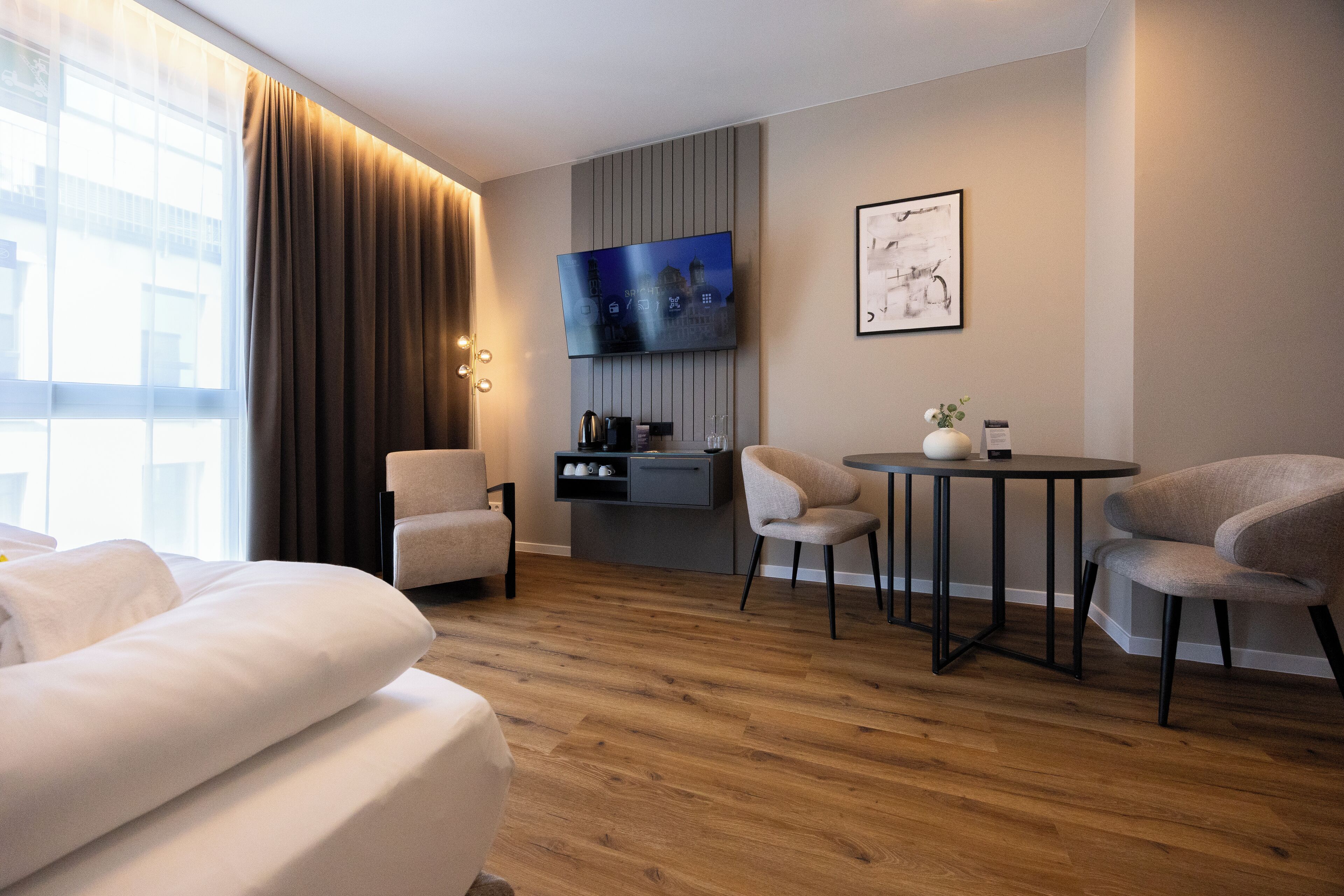 A modern hotel room with a large bed, two armchairs, a small round table, a TV mounted on the wall, and a kitchen area with a coffee machine.