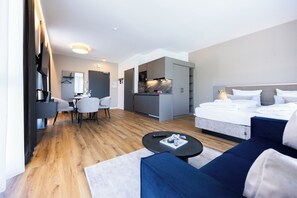 A modern hotel room with a wooden floor, a dining area with a table and chairs, a kitchenette, and a large bed with white bedding.