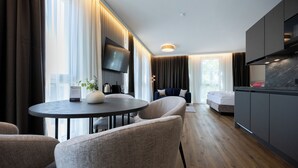 Moderni hotellihuone, jossa on ruokailutila, sohva, seinälle asennettu televisio ja pieni keittiö.