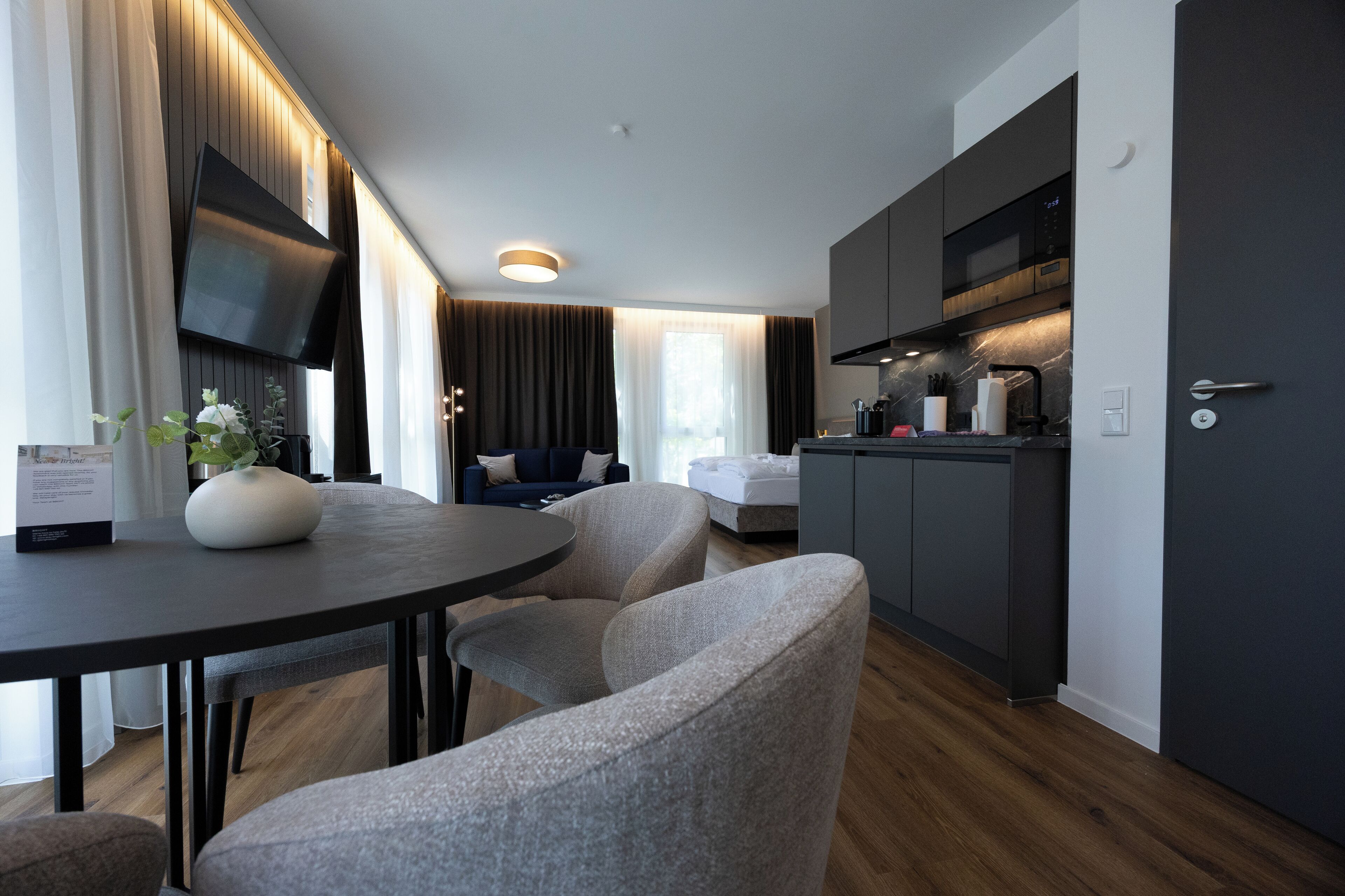 A modern hotel room with a dining area, a sofa, a TV mounted on the wall, and a kitchen area.