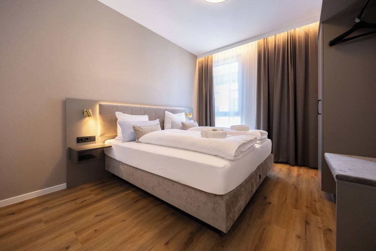 Een moderne hotelkamer met een groot bed, een nachtkastje met een lamp en uitzicht op de stad door grote ramen.
