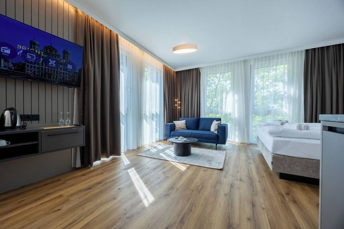 Een moderne hotelkamer met een houten vloer, een blauwe bank, een bed, een aan de muur bevestigde televisie en een bureau met een waterkoker.