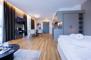 A modern hotel room with a wooden floor, a bed with white linens, a desk with a chair, a television, and a wardrobe.