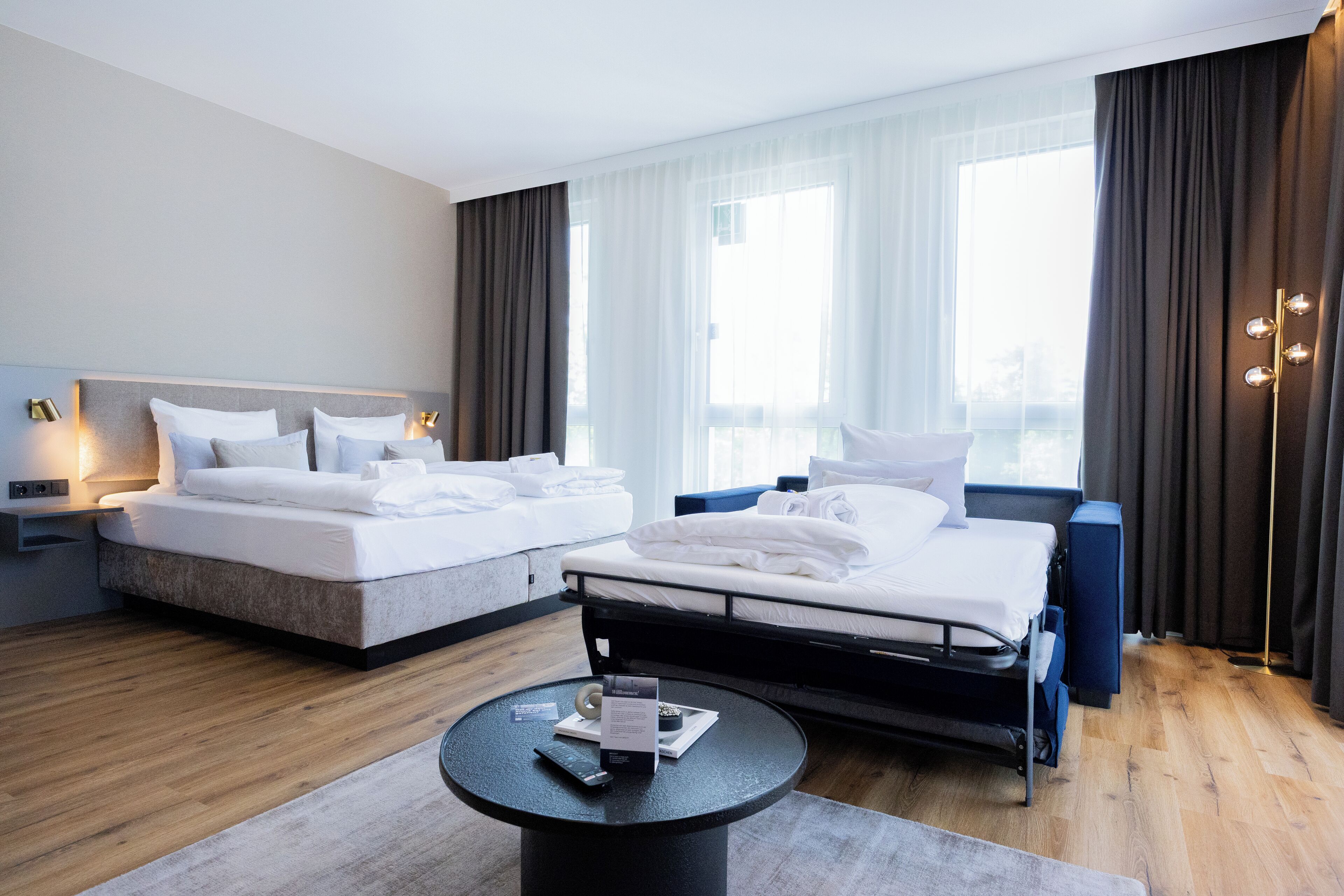 Una habitación de hotel moderna con dos camas, una mesita redonda y vistas de la ciudad a través de amplios ventanales.