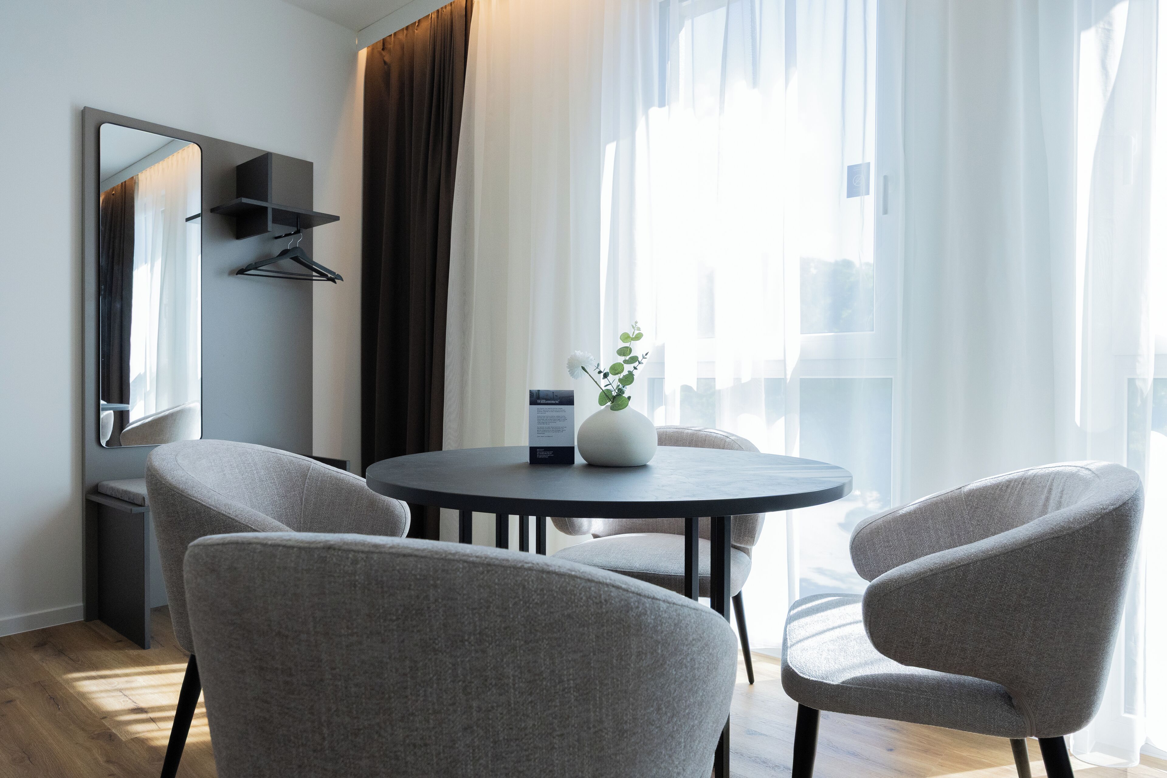 A modern hotel room with a round dining table, four grey upholstered chairs, a large mirror, a small white vase with a plant, and a dark-colored sofa.
