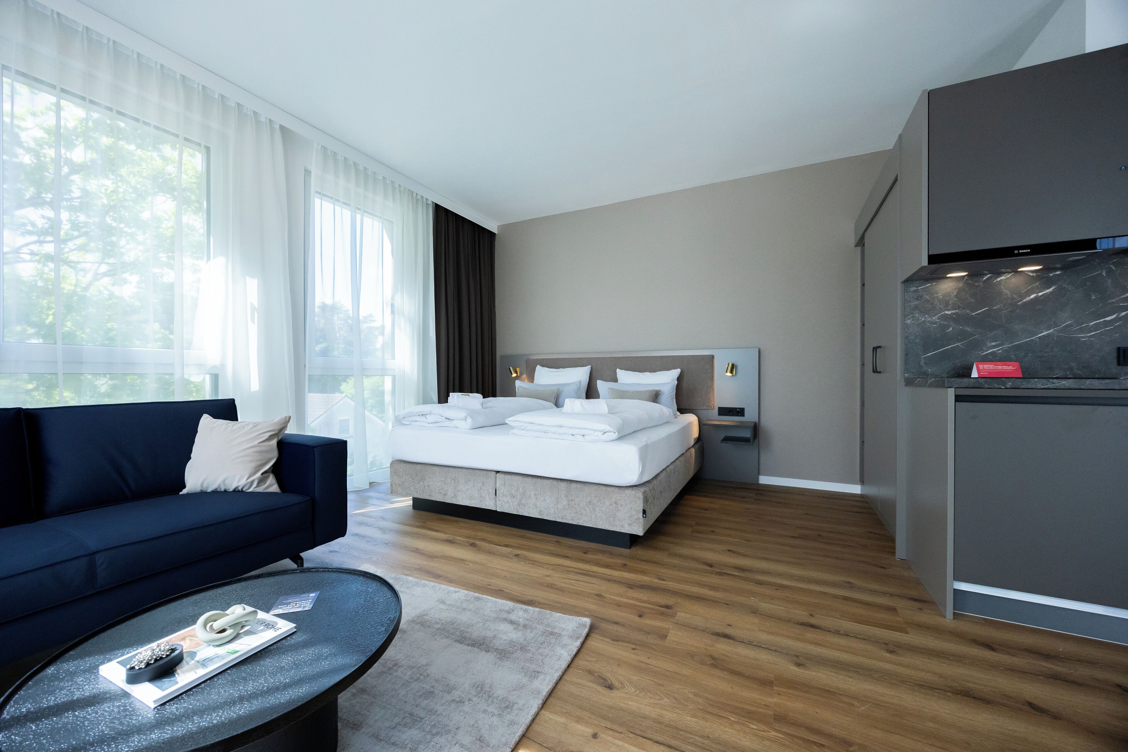 Ein modernes Hotelzimmer mit einem großen Bett, einem dunkelblauen Sofa, einem kleinen runden Tisch und einer Küchenzeile.
