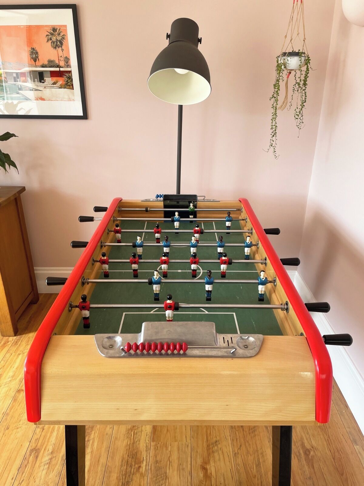 Een tafelvoetbaltafel met een rode rand, een zwarte lamp en een ingelijst foto aan de muur.