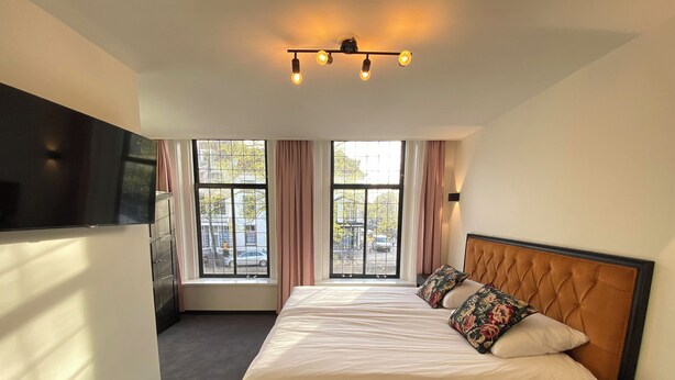 A hotel room with a large bed, a TV mounted on the wall, and two windows with curtains. - Hotel La Noire - Delft City Centre (Delft)