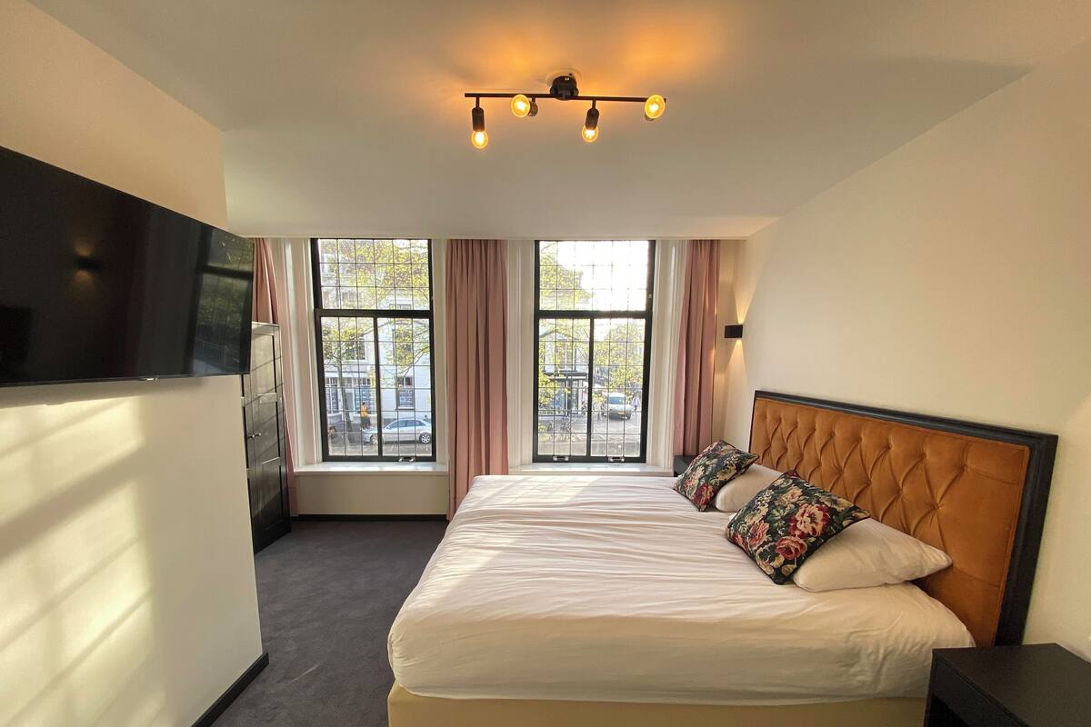 A hotel room with a large bed, a TV mounted on the wall, and two windows with curtains.