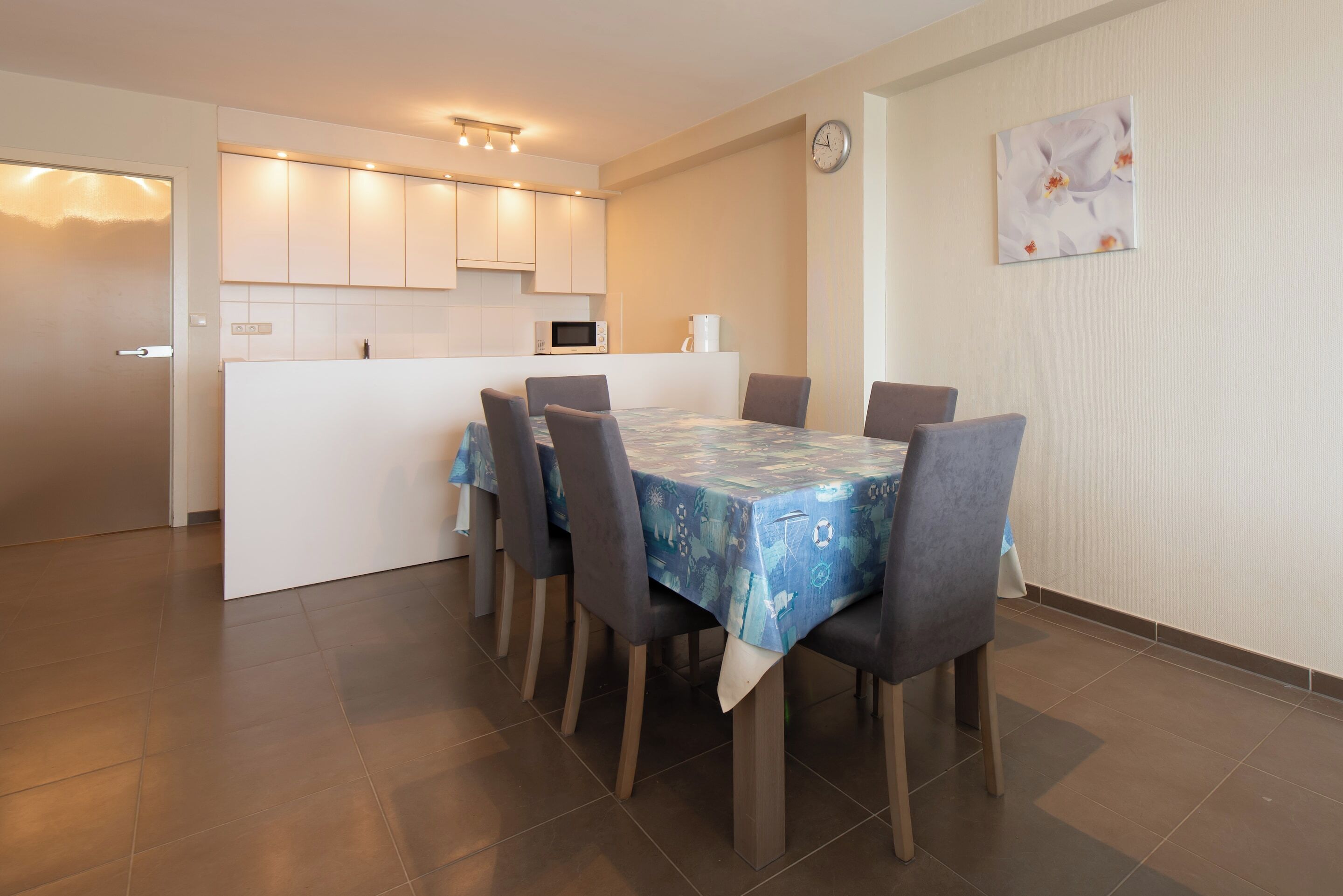 A modern kitchen with white cabinets, a microwave, and a coffee maker. There is a dining table with chairs and a clock on the wall.