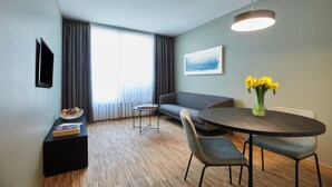 A modern hotel room with a dining area, a sofa, a TV mounted on the wall, and a window with curtains.