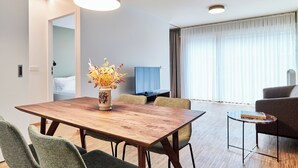 Une chambre d’hôtel moderne avec une table à manger en bois, des chaises recouvertes d’un tissu vert, un vase avec des fleurs séchées, un téléviseur à écran plat, un canapé et de grandes fenêtres avec des rideaux.