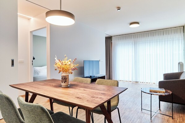 Una habitación de hotel moderna con una mesa de comedor de madera, sillas tapizadas en verde, un jarrón con flores secas, un televisor de pantalla plana, un sofá y ventanas amplias con cortinas.