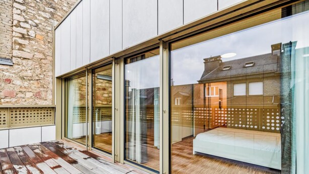 Une chambre d’hôtel moderne dotée de grandes portes-fenêtres coulissantes, d’une terrasse en bois et offrant une vue sur un bâtiment traditionnel.