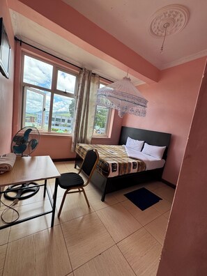 Un dormitorio con una cama, un escritorio, una silla, un ventilador de pie y una ventana con vistas a edificios y árboles.