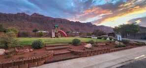 A playground with a slide and climbing structures, surrounded by landscaped gardens and a view of a mountain range.