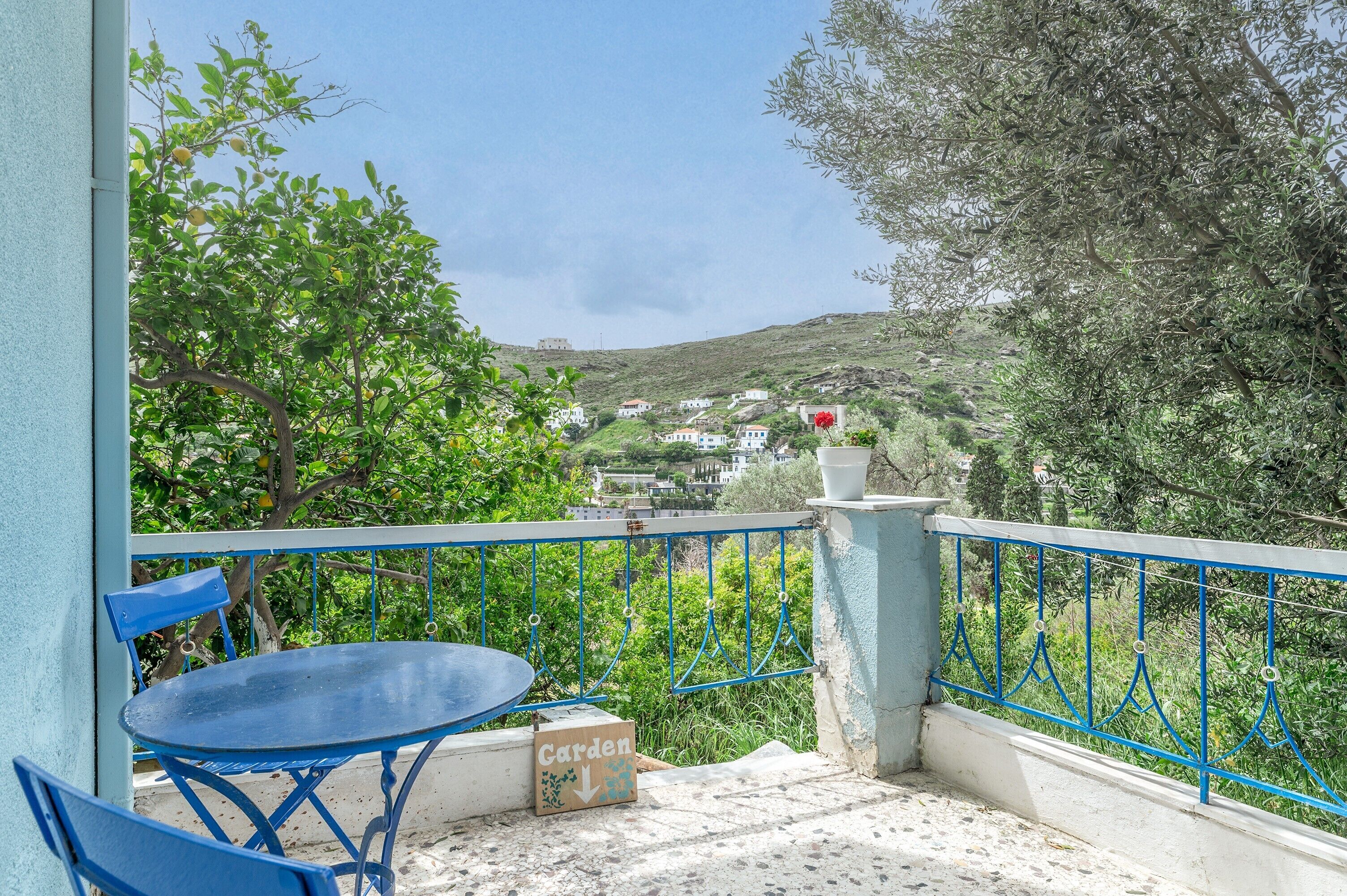 Uma varanda com mesa e cadeiras, um letreiro com a palavra 'Garden' e vista para uma encosta com edifícios.