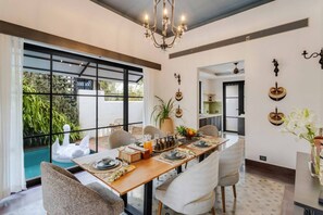 A dining area with a long table set for four, surrounded by chairs, with a view of greenery outside through large windows. - Lux 3BR Villa in Siolim 4km to Beach Wadi House (Siolim)