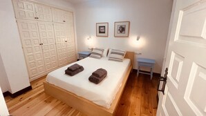 A bedroom with a wooden floor, a bed with pillows and folded towels, a wardrobe with a geometric pattern, and two framed pictures on the wall.