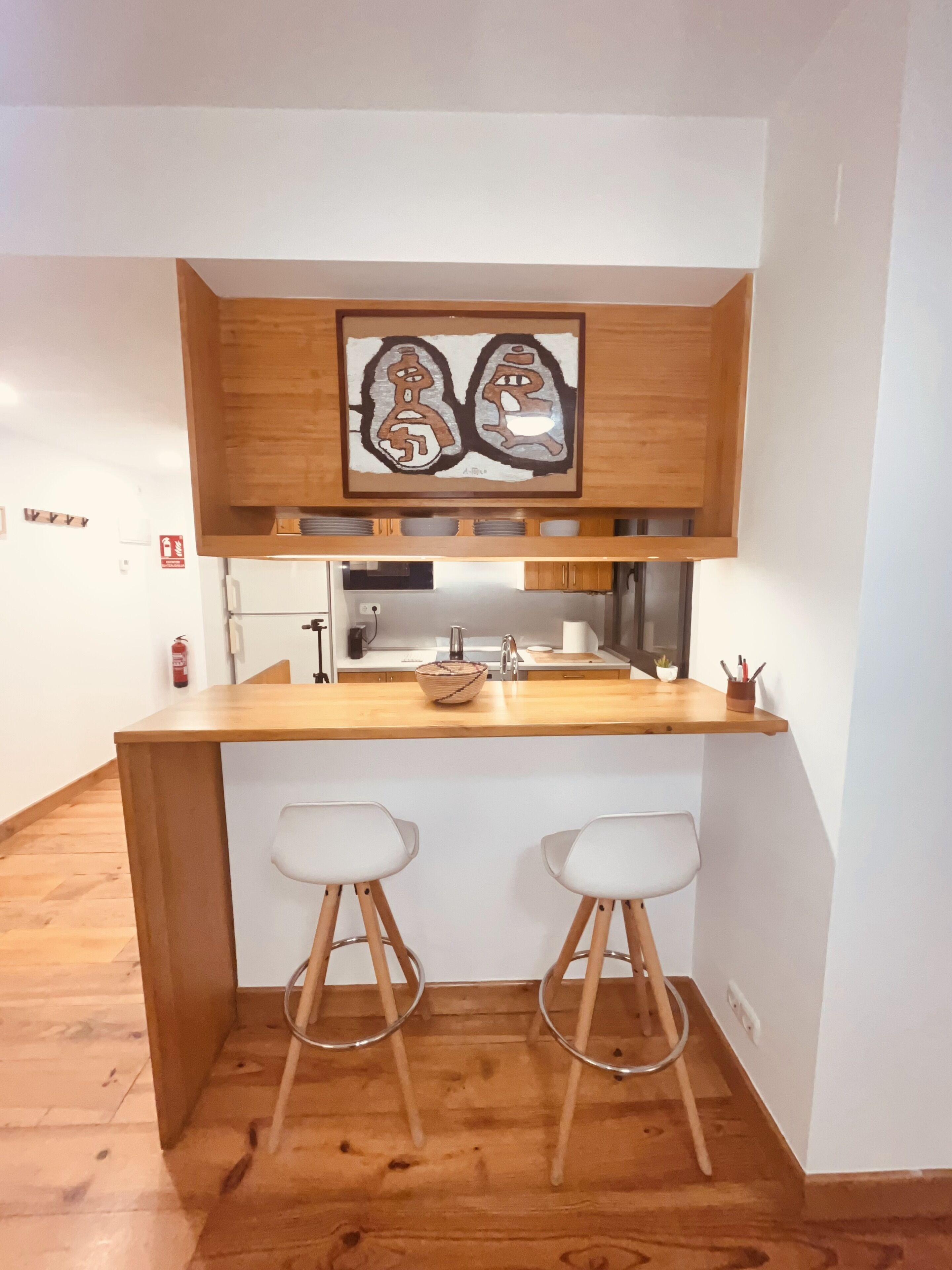 Une cuisine compacte dotée d’un plan de travail en bois, de deux tabourets de bar blancs et d’une œuvre d’art suspendue au-dessus de l’évier.