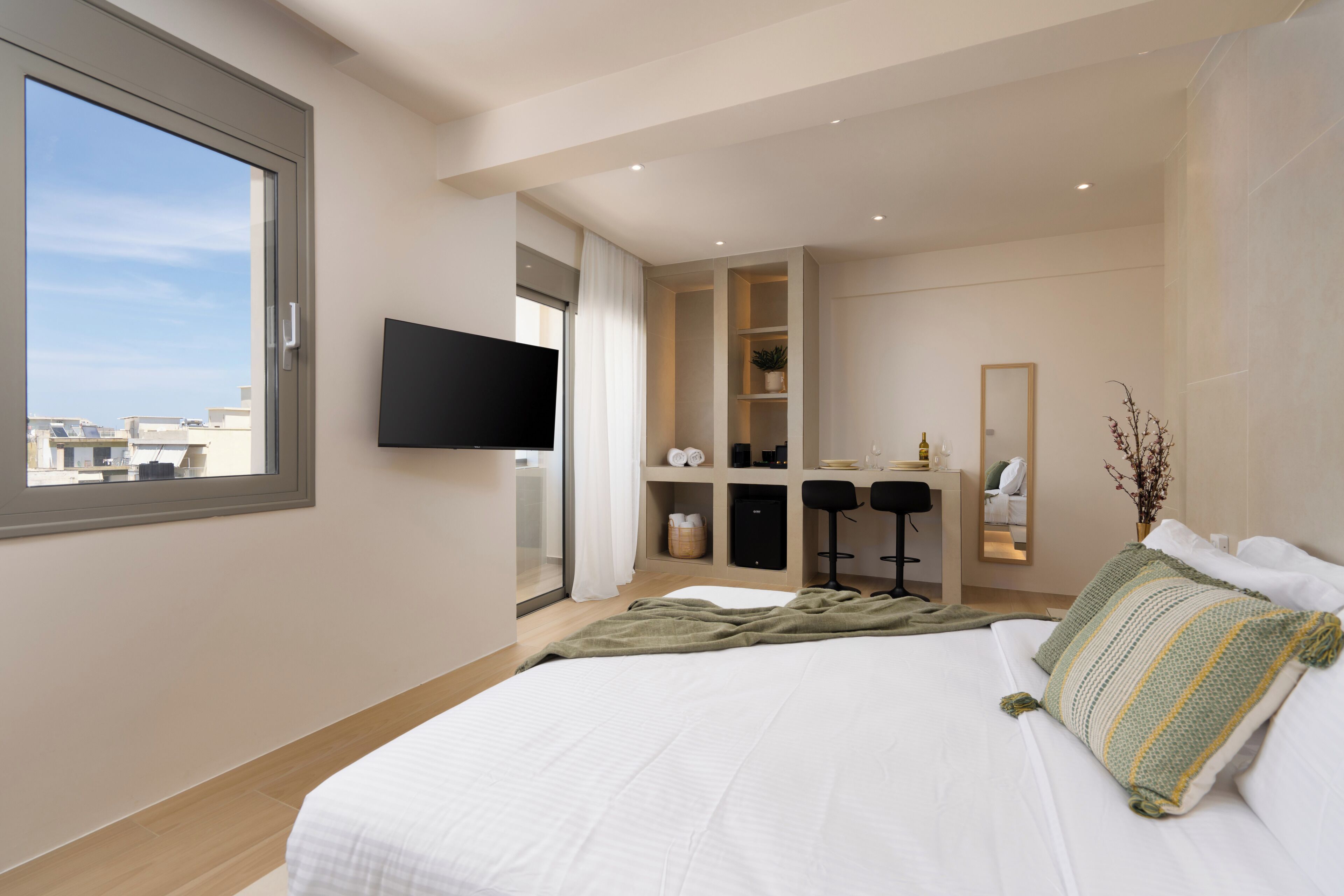 A modern hotel room with a large bed, a TV mounted on the wall, a desk with a chair, and a view of the city through the window.