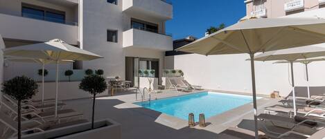 A pool area with sun loungers and umbrellas, surrounded by a white building with multiple balconies.