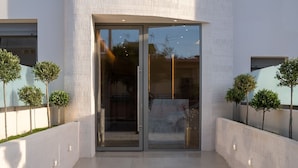 A modern building entrance with a glass door, a tiled pathway, and small trees along the sides. - Selected Suites  (Chania)
