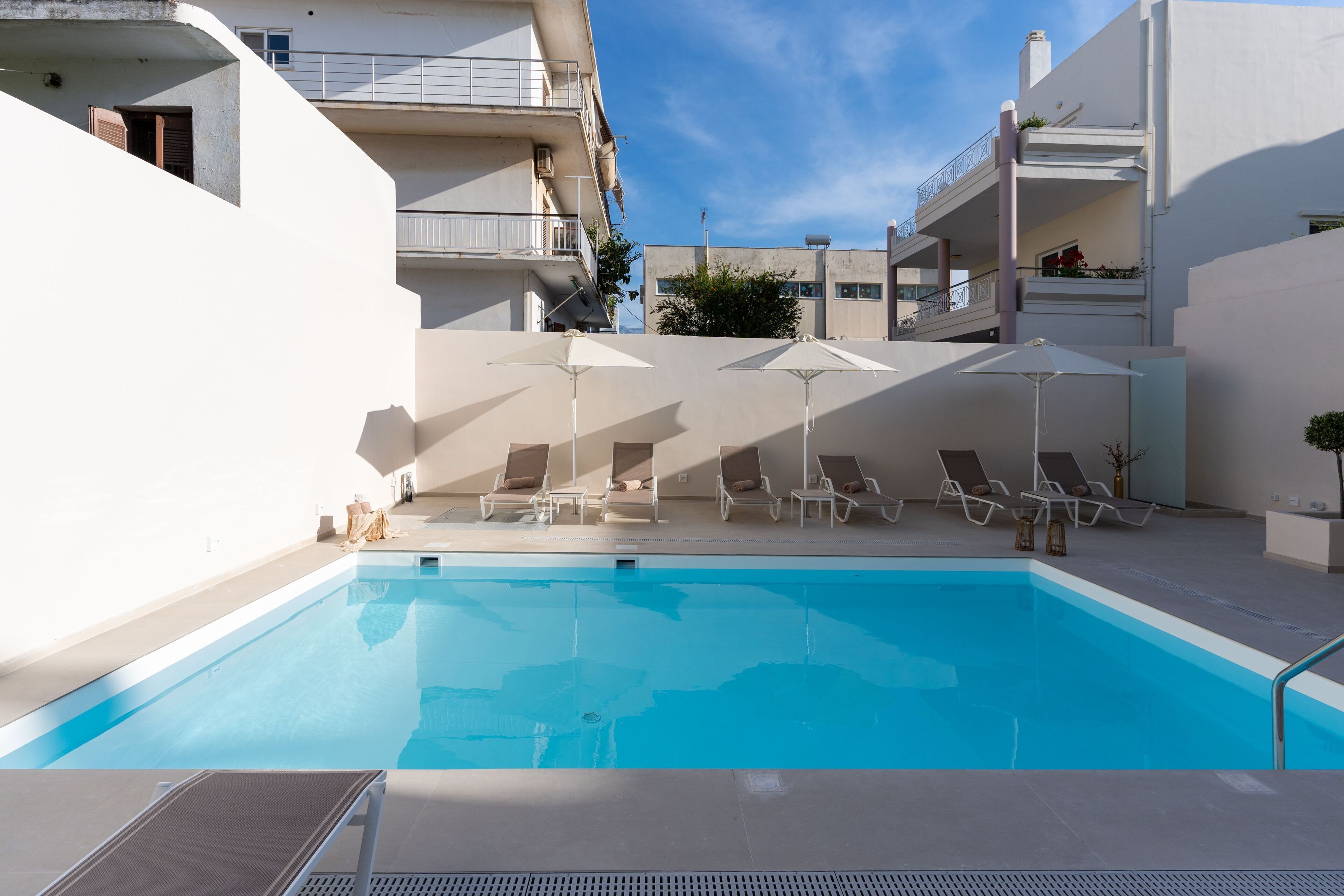 Une piscine entourée de bâtiments blancs dotés de balcons, avec des chaises longues et des parasols à proximité.