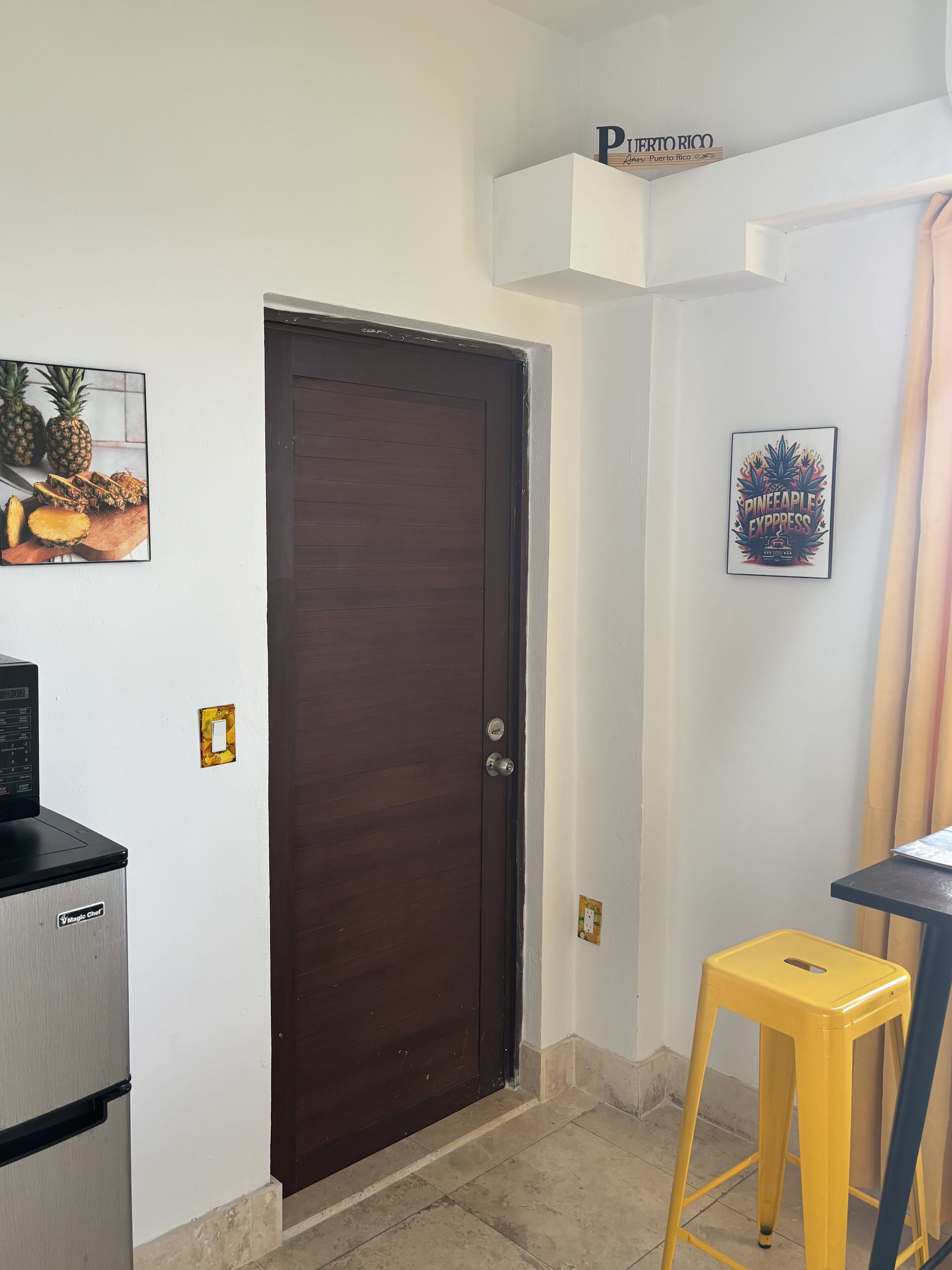 A closed dark wooden door, a small refrigerator, a yellow stool, and two framed pictures on the walls.