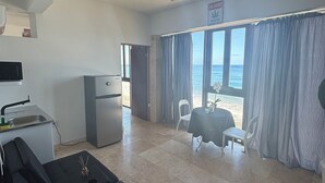 A compact living space with a kitchenette, a sofa, a dining area with a table and chairs, and a view of the sea through large windows. - BednBong Experience Stay (Arecibo)