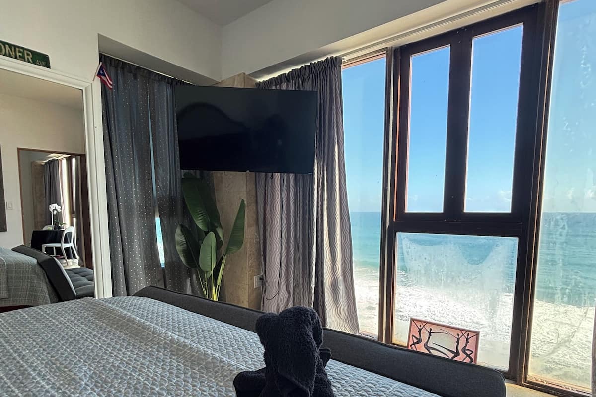A hotel room with a bed, a TV, a desk, and a large window overlooking the beach.