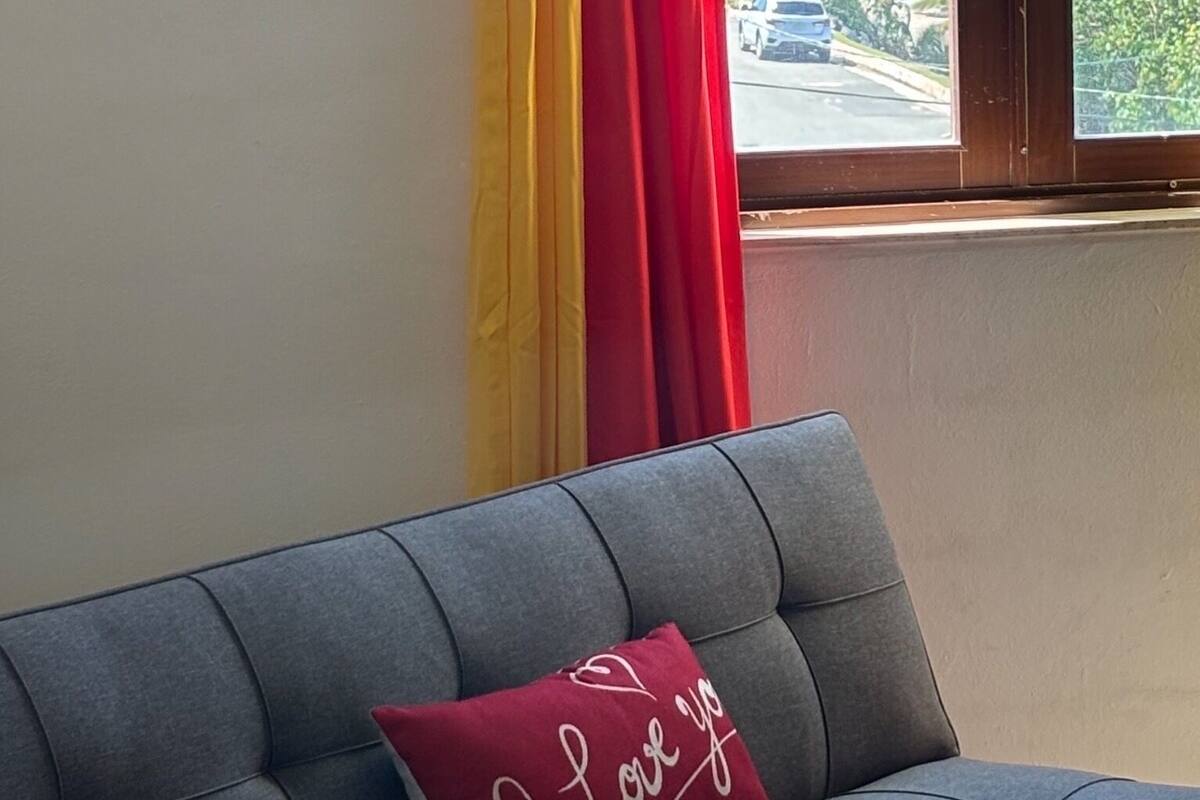 A room with a grey sofa, a red cushion with "I Love You" text, and a window with a view of the ocean and palm trees.