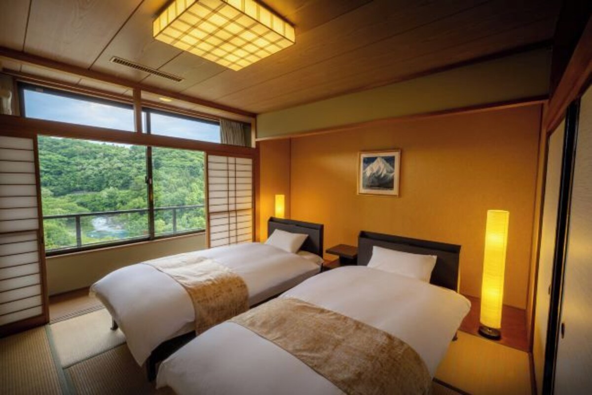 A hotel room with two beds, a large window, and a view of greenery.