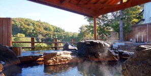Un onsen japonais traditionnel, avec des bains d’eau chaude, entouré de constructions en bois et surplombant une colline boisée.