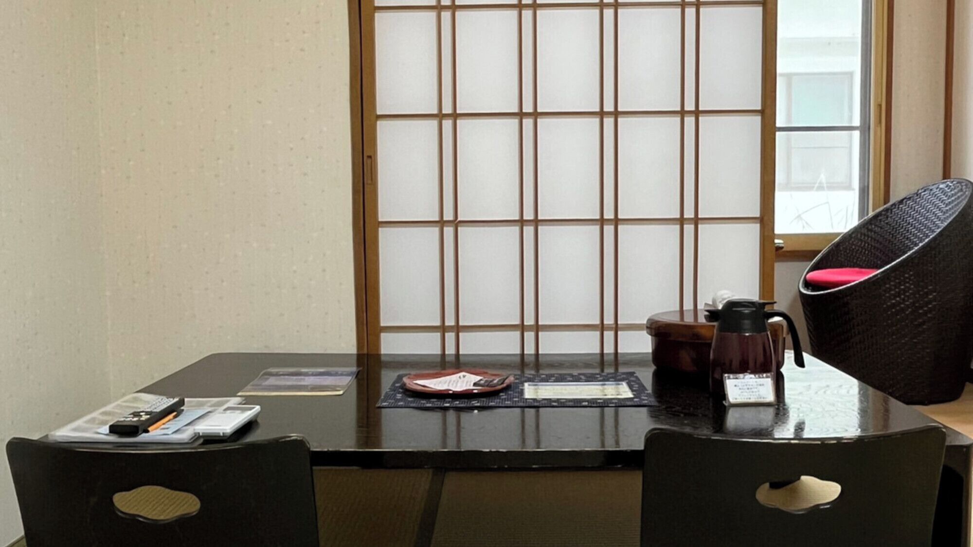Une pièce de style japonais traditionnel, avec un sol recouvert de tatamis, une table basse, deux chaises noires avec des coussins, un paravent shoji et une fenêtre donnant sur un paysage.