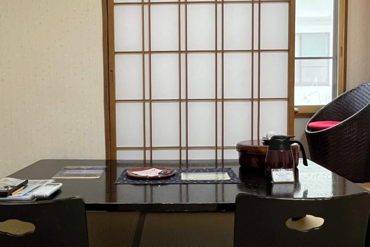 Une pièce de style japonais traditionnel, avec un sol recouvert de tatamis, une table basse, deux chaises noires avec des coussins, un paravent shoji et une fenêtre donnant sur un paysage.