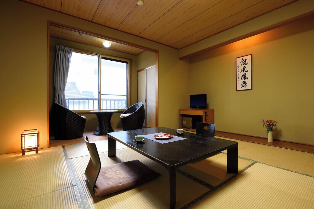 Une pièce de style japonais traditionnel, avec un sol recouvert de tatamis, une petite table basse avec un coussin, un téléviseur posé sur une étagère et une porte coulissante donnant sur un balcon.