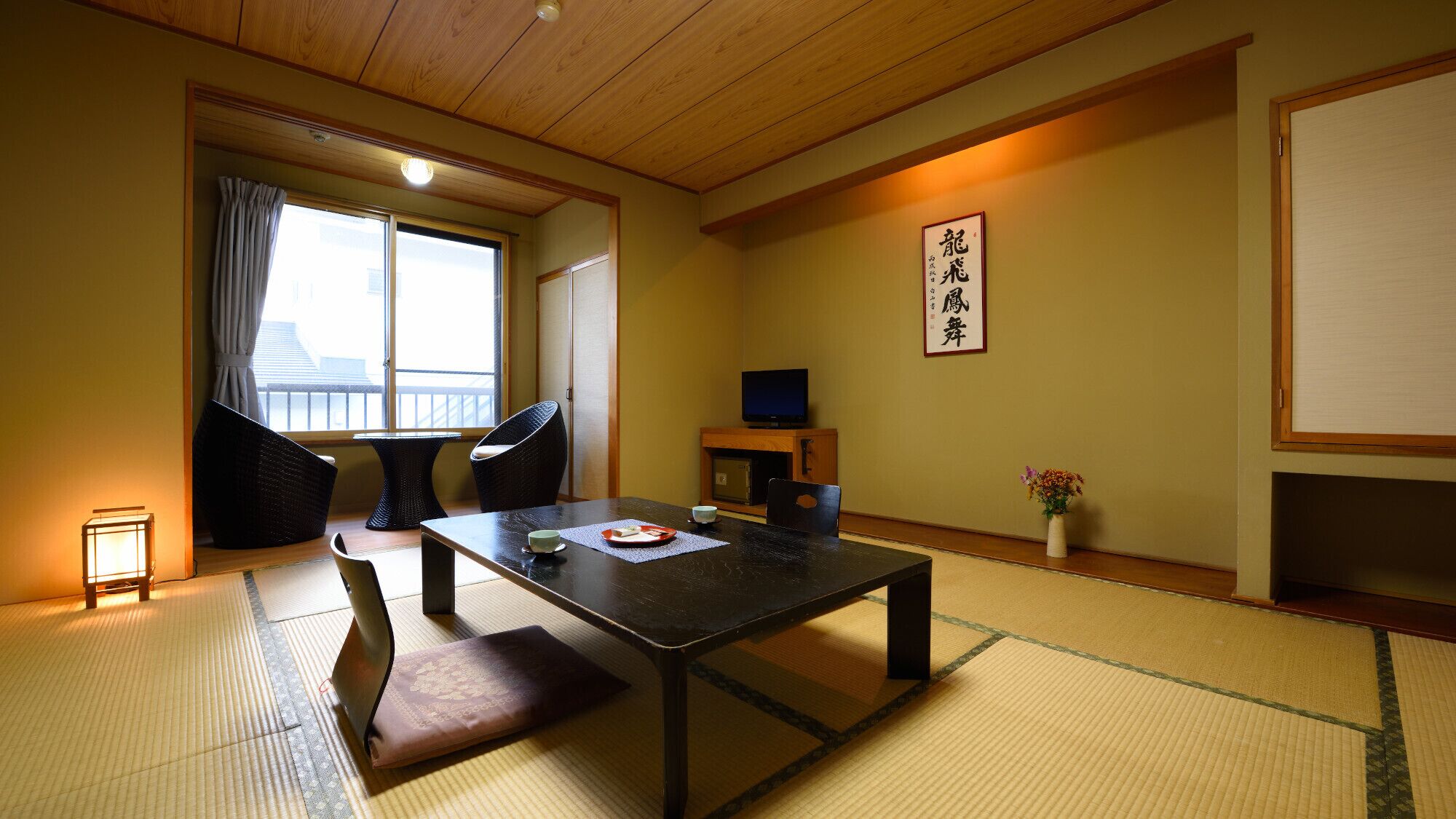 Une pièce de style japonais traditionnel, avec un sol recouvert de tatamis, une petite table avec un coussin, un téléviseur et une porte coulissante donnant sur un balcon.