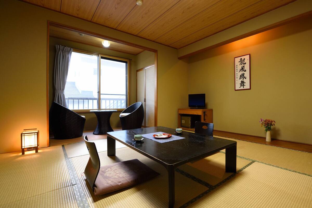 Une pièce de style japonais traditionnel, avec un sol recouvert de tatamis, une petite table avec un coussin, un téléviseur et une porte coulissante donnant sur un balcon.