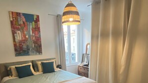 Une chambre avec un grand lit, un luminaire suspendu, un tableau au mur et une fenêtre avec des rideaux.