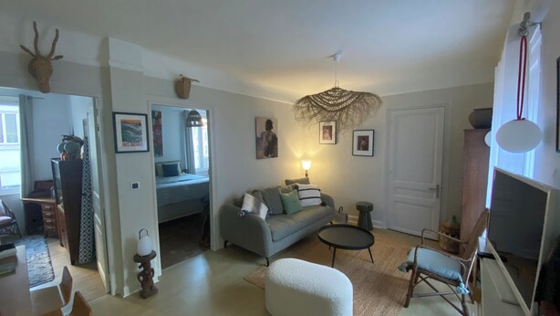 Un salon avec un canapé, une petite table ronde, un fauteuil et une lampe. On aperçoit une porte et une fenêtre au fond.