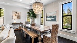 Un comedor con mesa de madera, banco y silla de mimbre, rodeado de ventanas amplias y obras de arte.